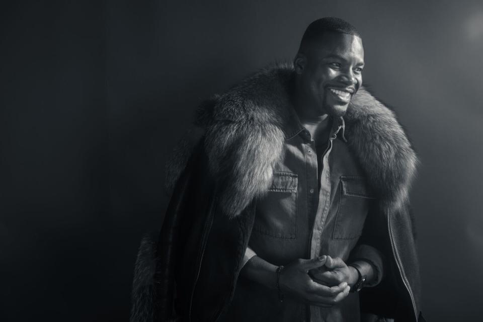 A man with a fur lines coat over his shoulders smiles.