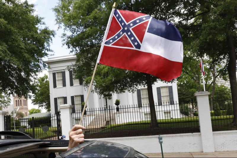 In this April 25, 2020 photograph, a small Mississippi state flag is held by a participant during a drive-by “re-open Mississippi” protest past the Governor’s Mansion, in the background, in Jackson, Miss. (AP Photo/Rogelio V. Solis)