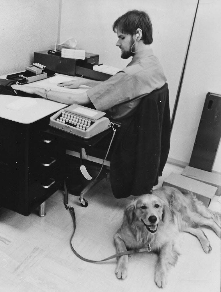 John Boyer works at his desk while his guide dog, Sugar, waits nearby.