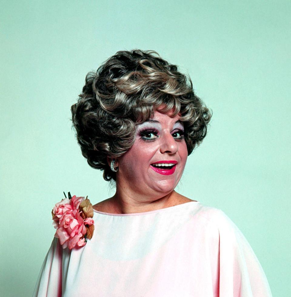 A woman with curly hair styled in an updo, wearing earrings and a white dress with a flower accessory, smiles at the camera