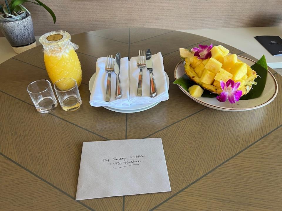 A table with pineapple juice in a pineapple-shaped carafe, plates with cutlery and napkins, and a pineapple shell with chopped pineapple and flowers.