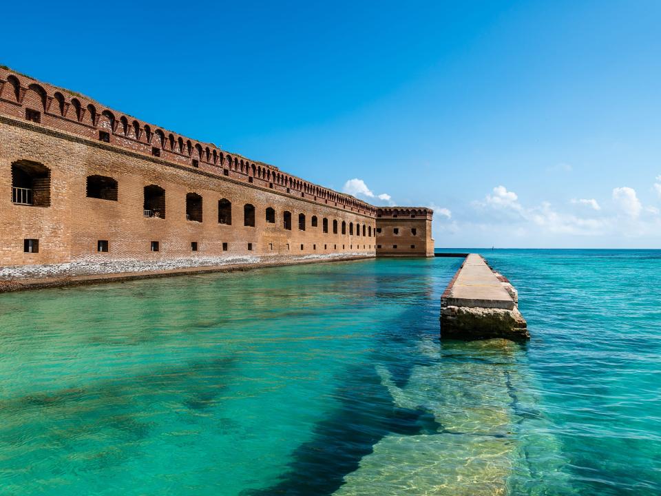 On the left is a long brick building with openings for windows. On the right is beautiful turquoise water and a brick wall in the water.