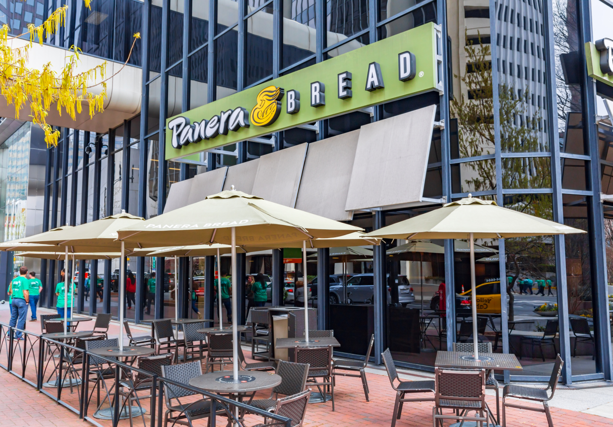 Front exterior Panera Bread restaurant in Uptown Charlotte, North Carolina, with two rows of outside seating in front, panel windows building on a fall day, yellow leaves of a tree on the left