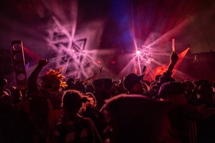 People dancing and holding lights at a crowded night festival, surrounded by colorful laser beams and smoke