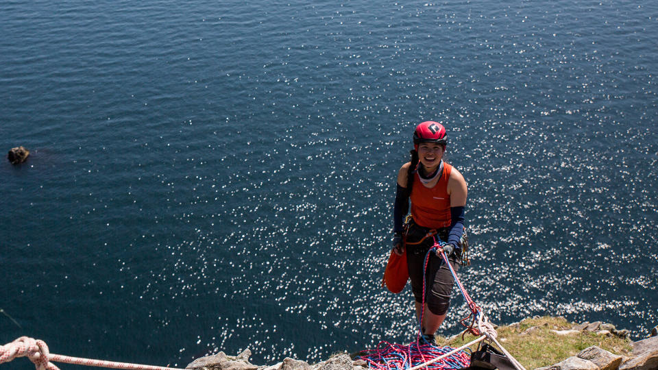 A woman stands on the edge of a grassy cliff, the sea behind her, holding on to the ropes attaching her. She is wearing a red helmet and a Sea to Summit Lightweight Dry Bag is attached to her waist.