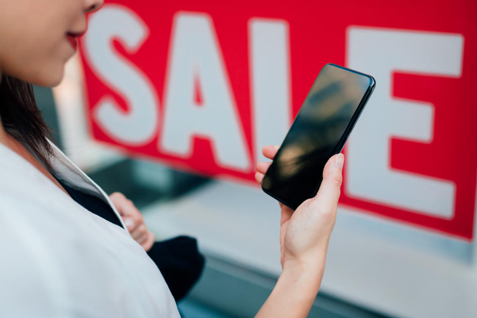 Person using smartphone, standing near a "SALE" sign, reflecting mobile shopping and promotions