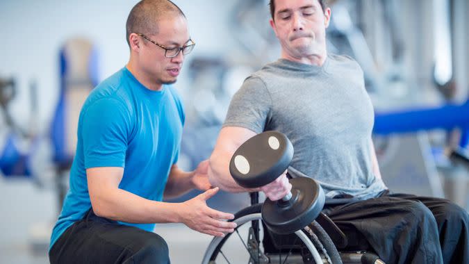 A man with a physical disability is sitting in a wheelchair and is working out at the gym with a recreational therapist