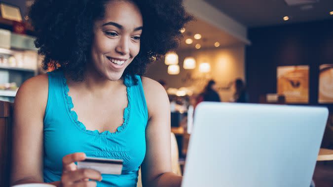 Smiling young African ethnicity woman sitting in cafe, holding a credit card and typing on a laptop.