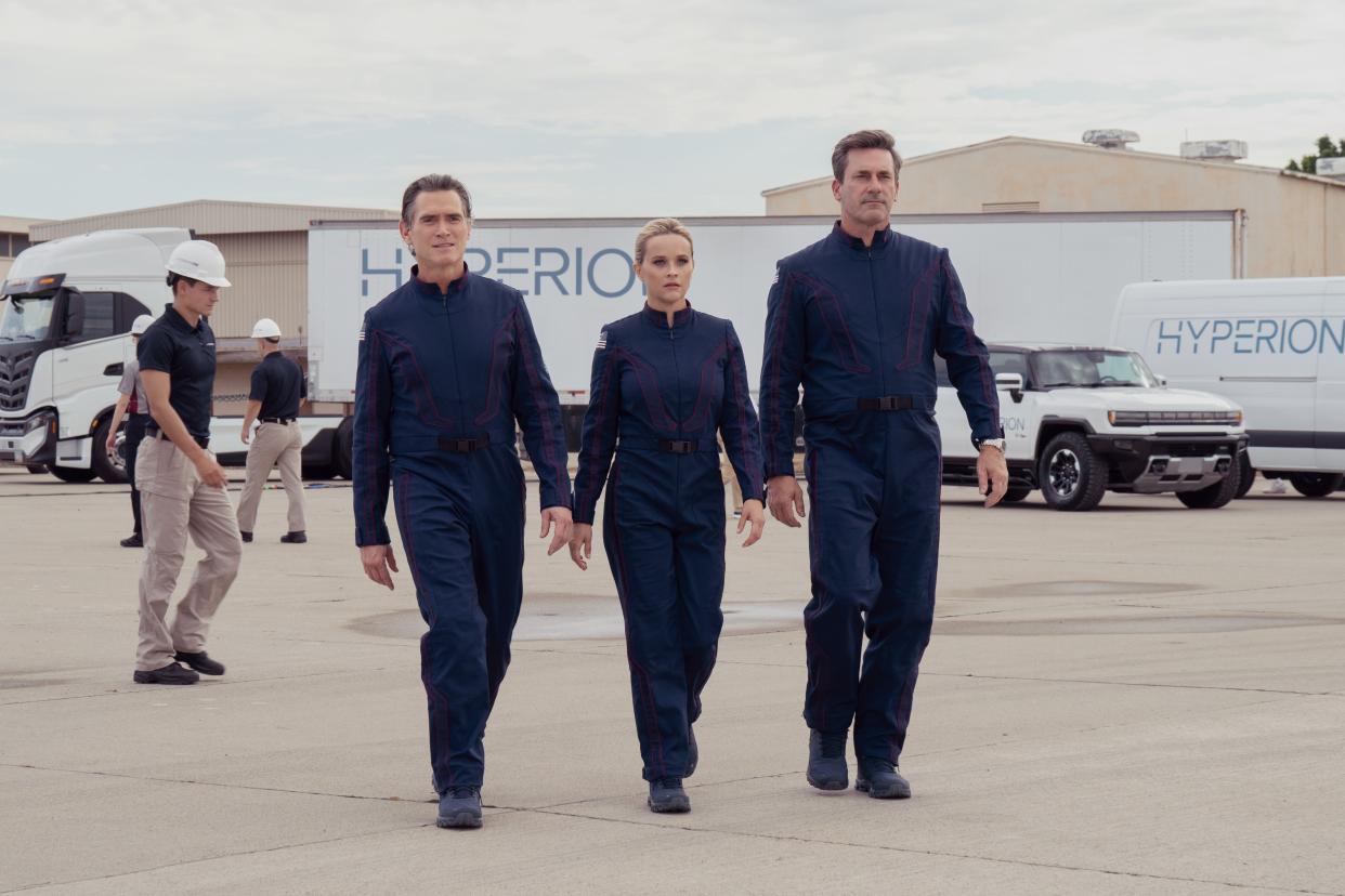 Billy Crudup, Reese Witherspoon, and Jon Hamm wearing spacesuits and walking on a lot.