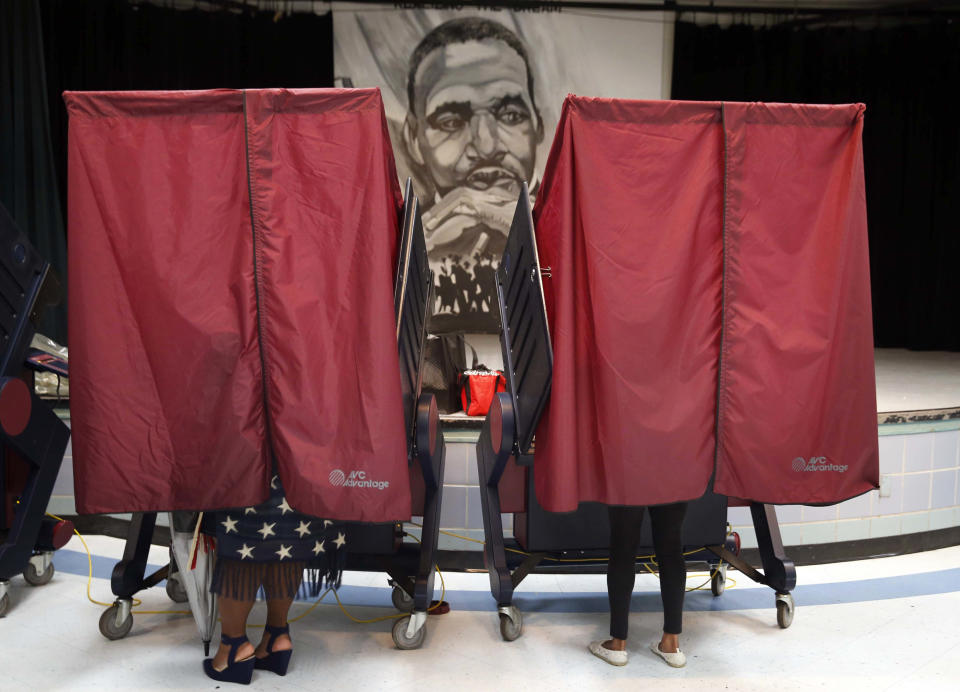 Voters behind the red curtains of two voting booths, one wearing a long navy skirt printed with stars, with an image of Martin Luther King, Jr., in the background.