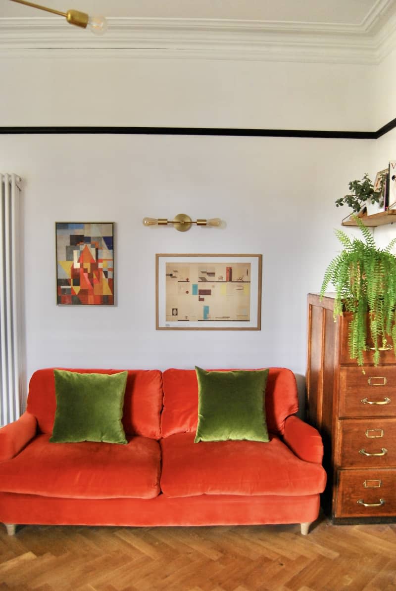 Red velvet love seat with green pillows next to wooden cabinets and walls with decorated artwork.