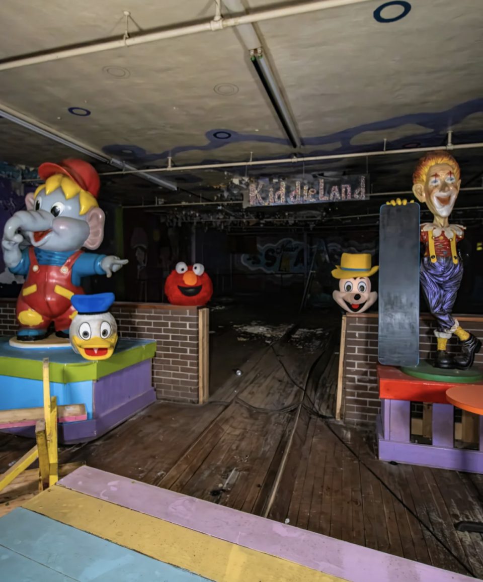 An indoor amusement area with large cartoon statues, including a clown, an elephant, a duck and a figure resembling Elmo, next to a "Children's Land" Sign