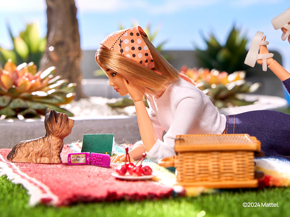 Se muestra una Barbie acostada en una mesa de picnic con un perro de plástico frente a ella y un teléfono rosa abierto de costado.