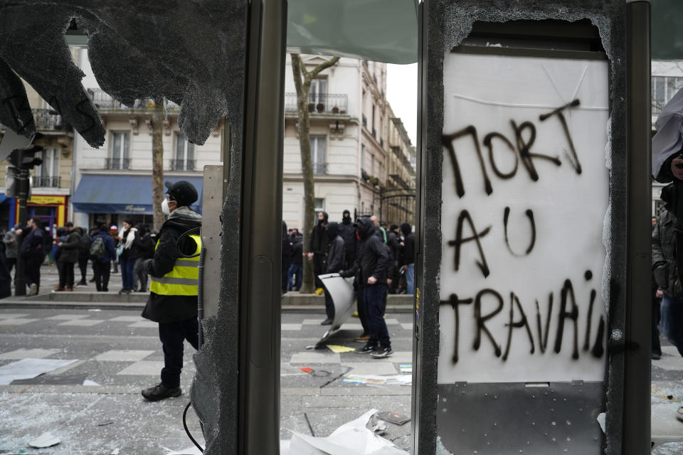 Youths stand by a destroyed bus stop reading "Death to work" during clashes with police officers during a demonstration, Tuesday, March 7, 2023 in Paris. Demonstrators were marching across France on Tuesday in a new round of protests and strikes against the government's plan to raise the retirement age to 64, in what unions hope to be their biggest show of force against the proposal. (AP Photo/Lewis Joly)