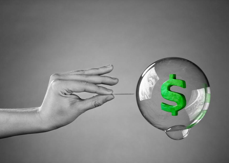 A person using a pin to pop a bubble containing a green dollar sign.