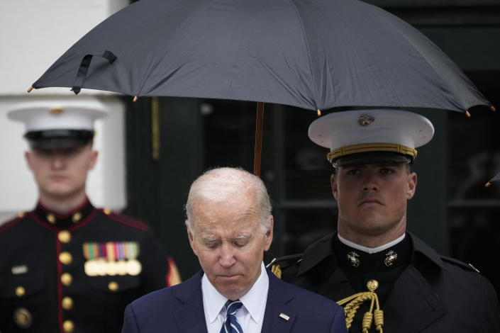 President Biden, with two Marines standing behind him, one holding an umbrella over Biden's head, looks down.