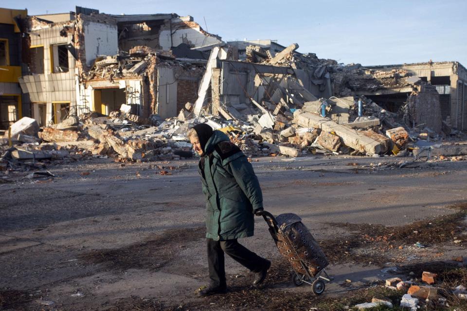 An elderly woman in a green coat pulls a trolley bag past a destroyed building in Bakhmut