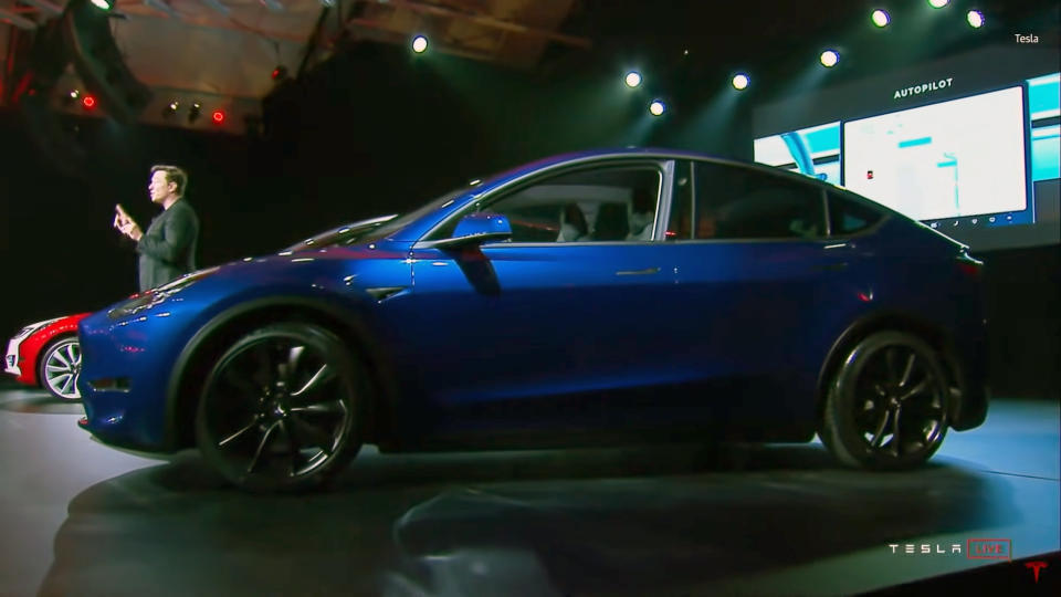 A blue Tesla Model Y electric vehicle sits on stage in a 2019 event. Elon Musk is seen behind it, speaking to an audience from a profile view relative to the camera.