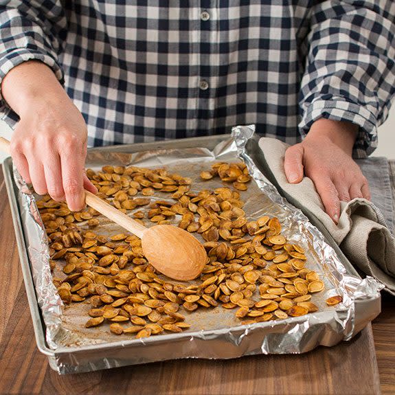 Person using a wooden spoon to stir perfectly roasted pumpkin seeds on a baking sheet lined with foil