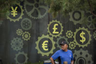 FILE - In this July 20, 2018, file photo, a deliveryman stands near a mural displaying Chinese yuan and other world currency symbols on the outside of a bank in Beijing. The official cost of the postponed Tokyo Olympics has increased by 22%, the local organizing committee said Tuesday, Dec. 22, 2020, in unveiling its new budget.(AP Photo/Mark Schiefelbein, File)