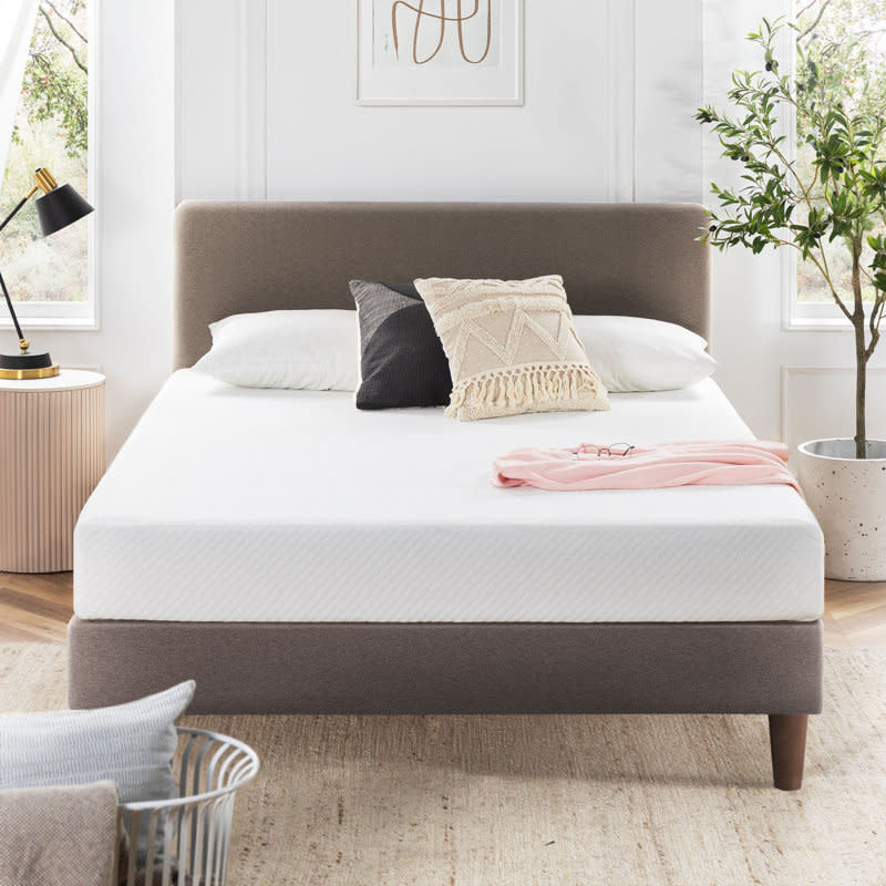 A minimalist bedroom features a neatly made bed with a light brown headboard, white mattress, and decorative pillows, set in a bright room with large windows, a plant, and modern decor