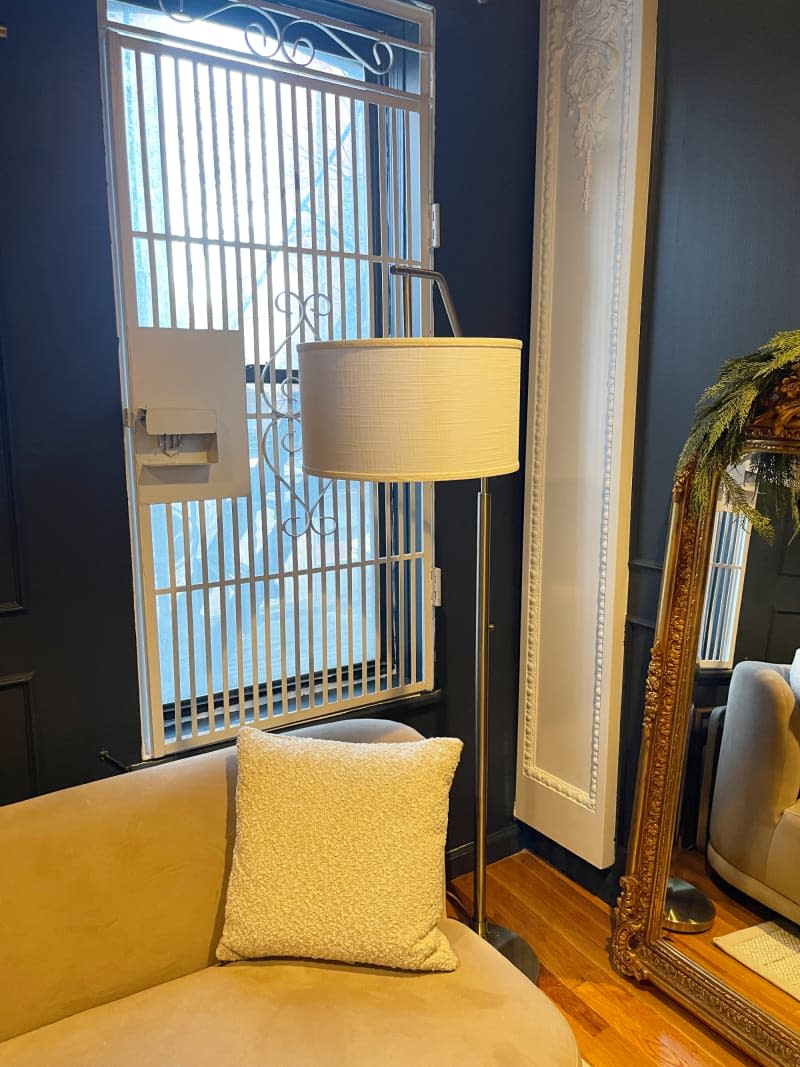 floor lamp with beige lamp shade in front of blue wall with gate on window and gold framed mirror