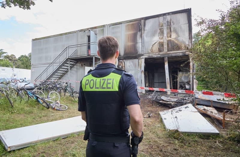 A police officer stands in front of sections of wall in front of accommodation containers following an explosion at a refugee shelter.  There is an explosion in a refugee shelter in Buchholz in der Nordheide.  The building is on fire.  One person dies and a police officer is seriously injured.  Georg Wendt/dpa