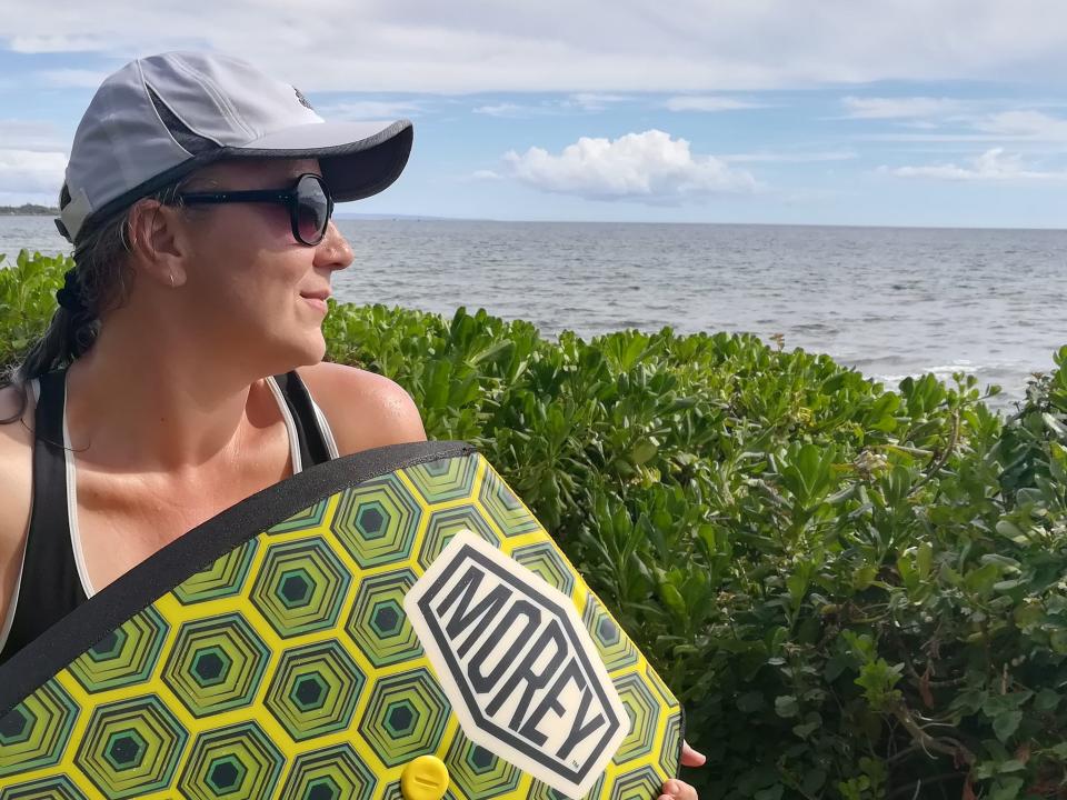 The author looking out to sea while wearing baseball cap and sunglasses with a green boogie board in her hands.