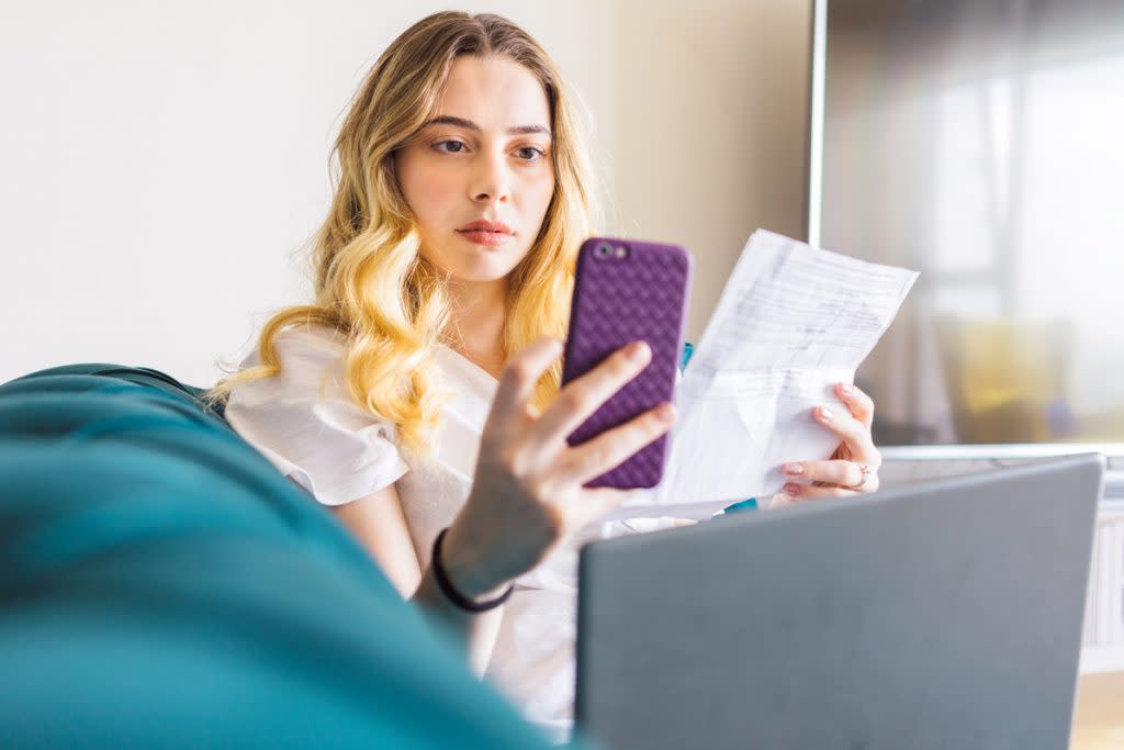 A woman tracks her spending while sitting on the couch with her phone, laptop and receipts. 