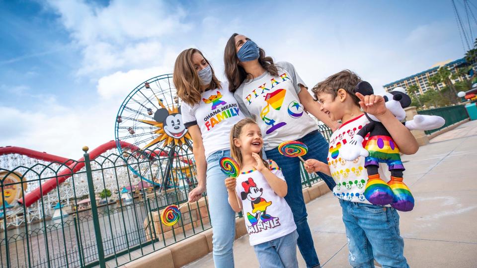 a family of four with two moms and two daughters stand at disney park wearing pride colored shirts 