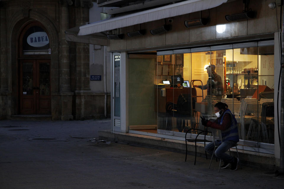 A man wearing a fask mask to protect againt coronavirus, handles his mobile telephone as he sits outside a shuttered cafe on the vacant Ledra Street in Nicosia, Cyprus, Sunday, April 26, 2020. Medical experts say the low number of new coronavirus infections is paving the way to start gradually lifting a strict day-at-home order that includes a night time curfew. (AP Photo/Petros Karadjias)