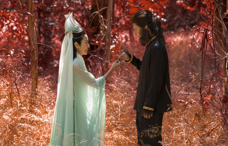 A woman in pale blue robes speaks to a boy wearing primate makeup in a forest full of red leaves; still from "American Born Chinese"