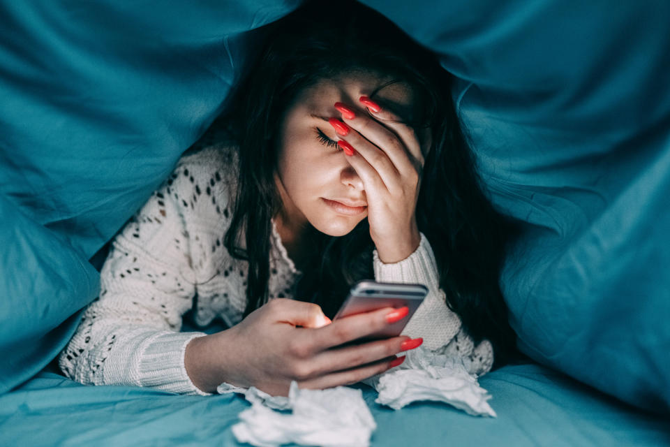 A woman on her phone under a blanket, with her hand on her forehead.