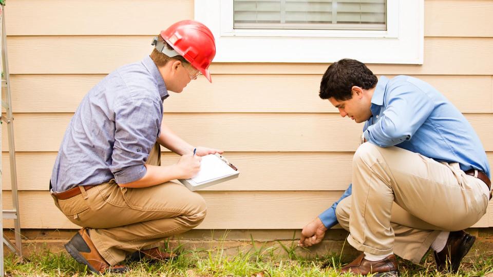 Repairmen, building inspectors, exterminators, engineers, insurance adjusters, or other blue collar workers examine a building/home's exterior wall and foundation.