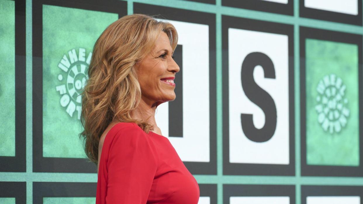 vanna white, seen in profile, smiles as she looks forward, she wears a red dress and stands in front of the wheel of fortune game board