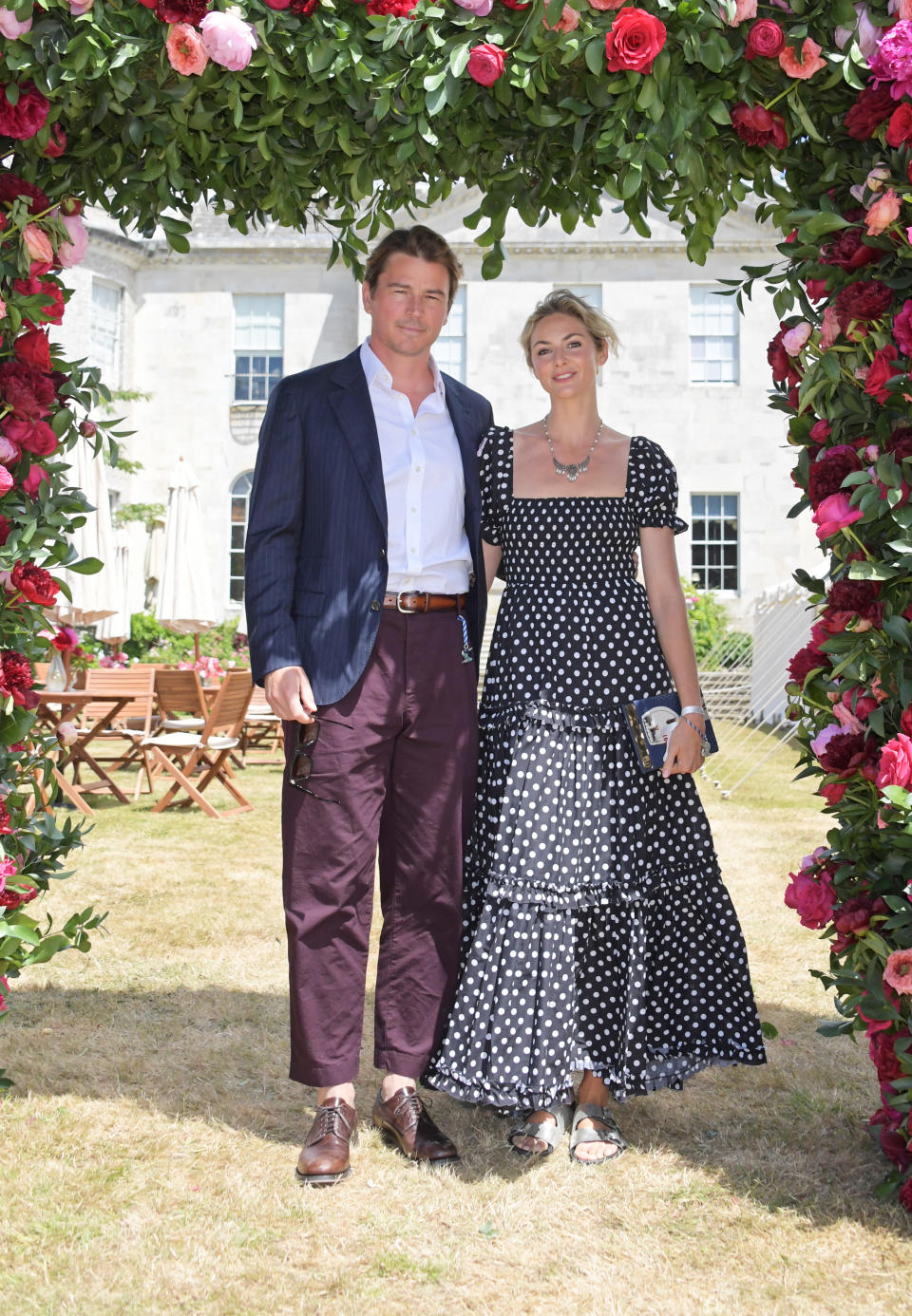 Josh Hartnett and Tamsin Egerton stand under a floral arch with out door dining and country manor in the background