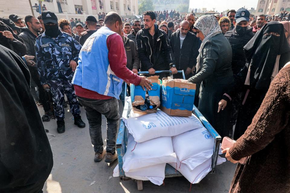 A man in a blue vest stands over a cart that has large white bags and cardboard boxes on it. Many men, including one in a blue camp uniform, surround him.