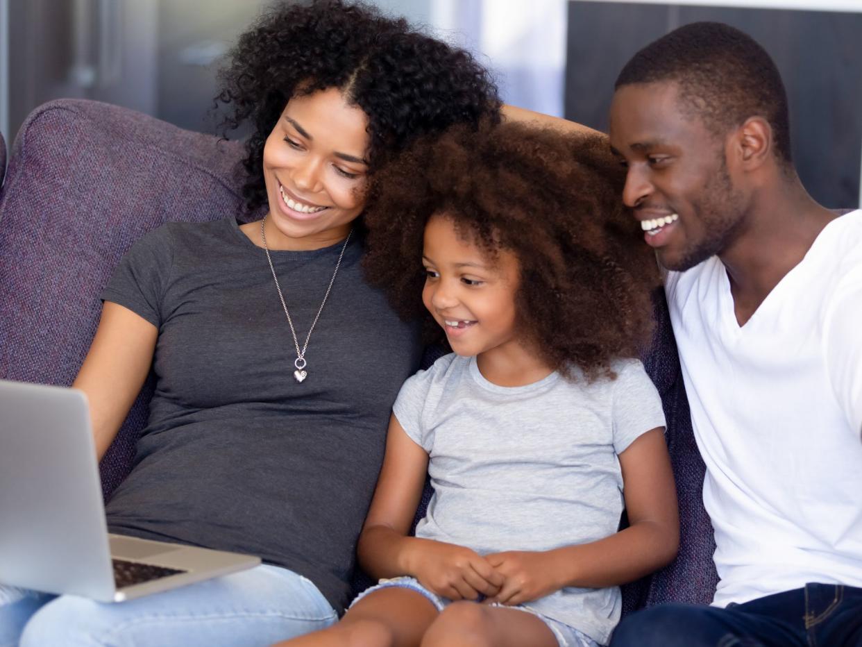 Smiling African American family using laptop together in living room, sitting on cozy sofa, looking at screen happy mother and father with little daughter watching comedy movie, making video call