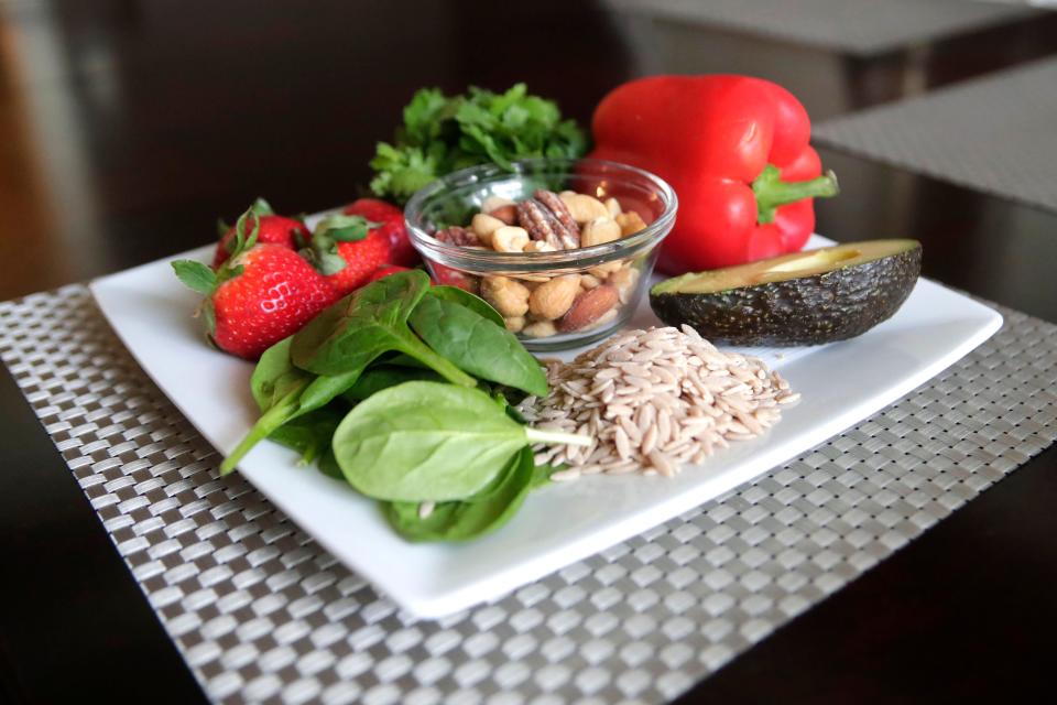 A heart-healthy plate of food is shown, including strawberries, cilantro, a red bell pepper, half an avocado, whole grains, spinach, and a bowl of mixed nuts.