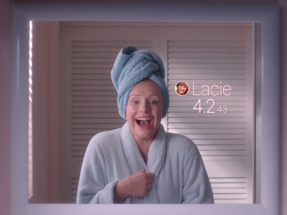 bryce dallas howard as lacie in black mirror, wearing a bathrobe and towel on her head. she's laughing widely while looking at herself in the mirror. to her right, there's a small photo of her, the name "lacie," and a 4.243 number