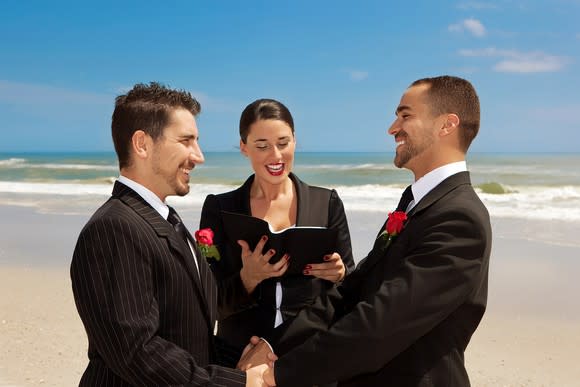 Two men in formal wear holding hands facing each other in front of an officiant holding a book, on a beach.