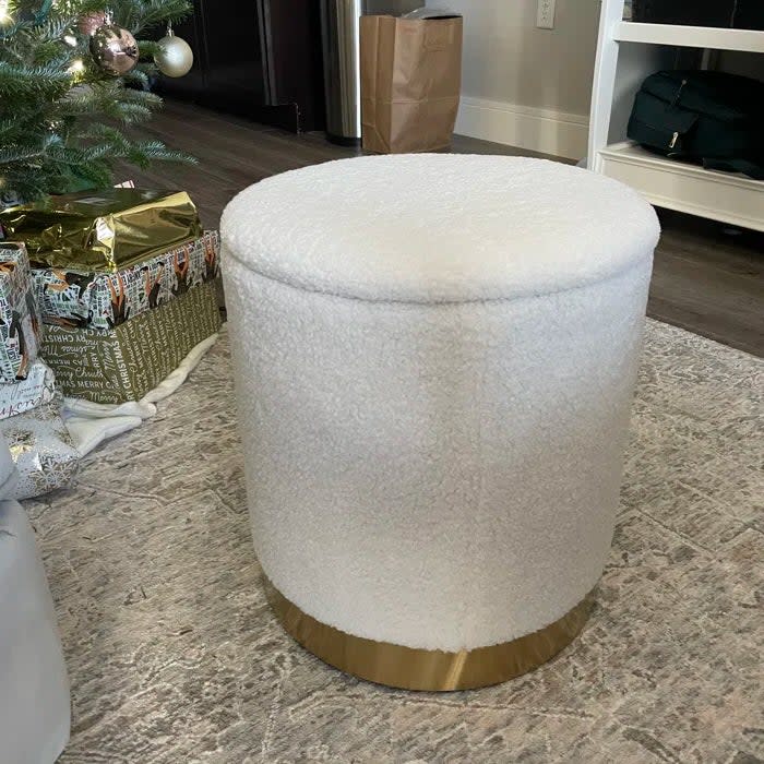 Reviewer's photo of the white, fluffy storage ottoman with a gold base, placed in a room with gifts and a tree in the background
