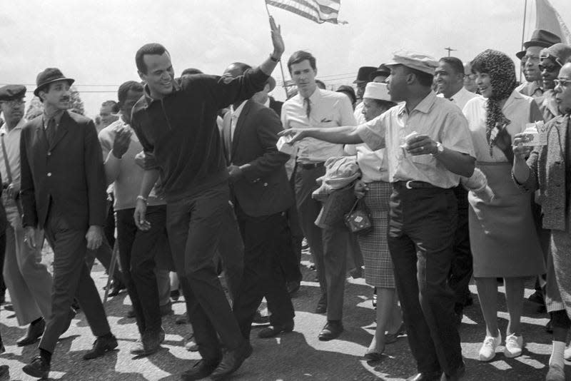 Harry Belafonte waves to Dr. Martin Luther King, Jr. (right) as he leaves the column of civil rights marchers here March 24th after walking with them for a short time. Belafonte is one of the show world celebrities who have traveled to Montgomery to entertain the Selma-to-Montgomery marchers on their final night encampment near the city limits of Montgomery. In the background (center) is movie star Tony Perkins, one of the luminaries taking part in the protest march.