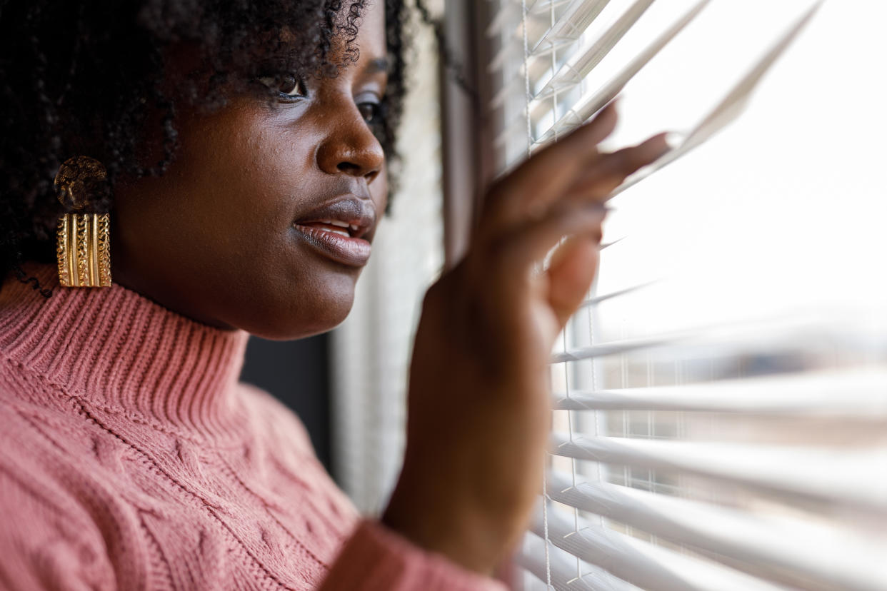 Portrait of young Black woman moving the blinds on the window, peeking outside, contemplating.