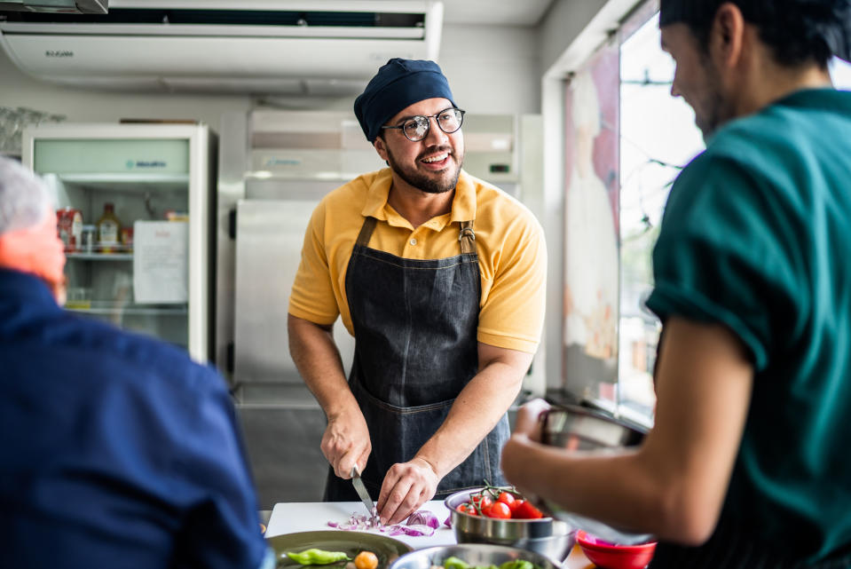 A man in a chef's apron chops onions in a kitchen while talking to another person