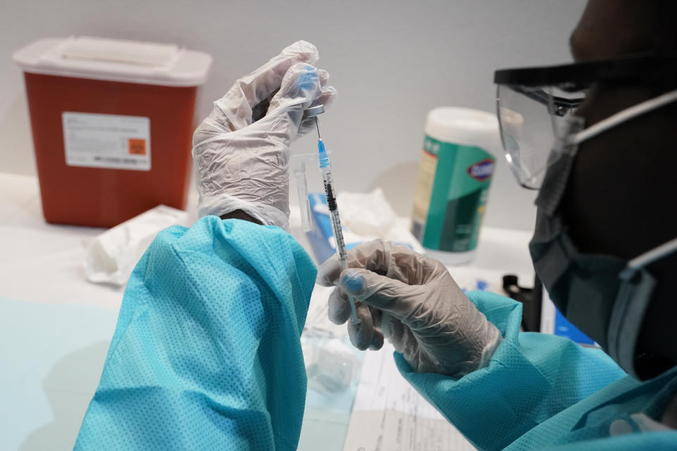 A health care worker wearing eye protection, a face mask and rubber gloves fills a syringe from a vial.