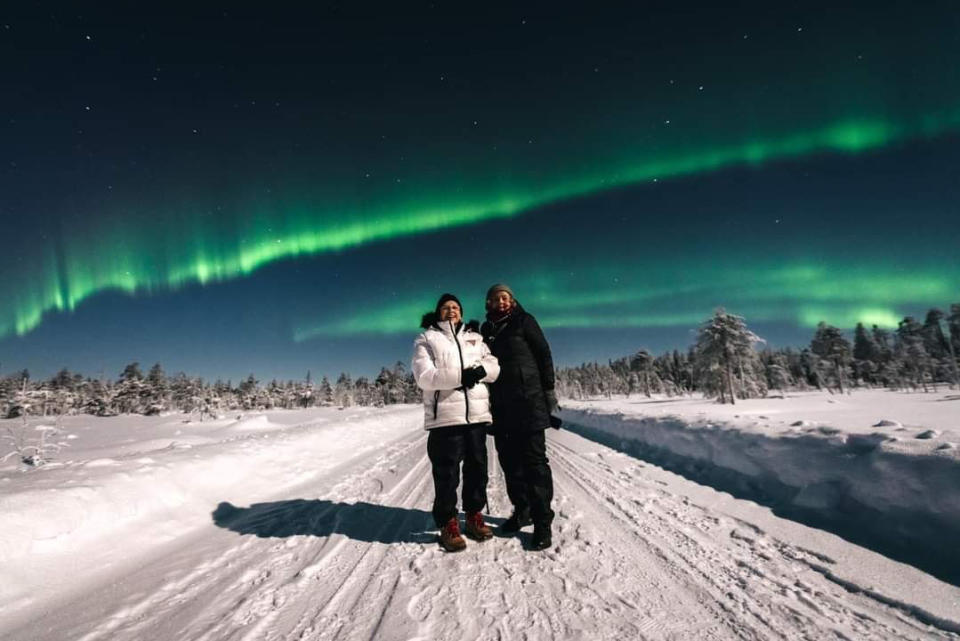 Two women in winter clothes on a snowy background stand in front of green glowing Northern Lights (Around the World at 80 / Facebook)