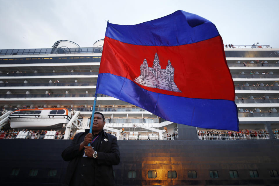 A Cambodian man holds his national flag in front of the MS Westerdam, owned by Holland America Line, dockked at the port of Sihanoukville, Cambodia, Friday, Feb. 14, 2020. Hundreds of cruise ship passengers long stranded at sea by virus fears cheered as they finally disembarked Friday and were welcomed to Cambodia. (AP Photo/Heng Sinith)