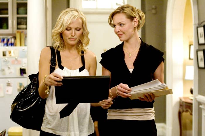 Malin Akerman and Katherine Heigl stand together, discussing something on a black frame, with papers in their hands in a scene from "27 Dresses"