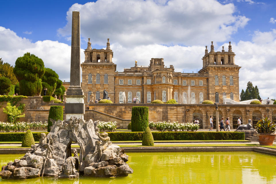 Woodstock, United Kingdom - June 27, 2015: Blenheim Palace, Woodstock, Oxfordshire, England. It is the principal residence of the dukes of Marlborough, and was built between 1705 and 1722. It is being used as a family home, mausoleum and national monument. The palace was also the birthplace of Sir Winston Churchill. Fountain, water, reflection, landscaped bushes and trees, sightseeing tourists and vivid blue sky with clouds are in the image.